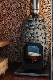 The HUUM Hive Flow LS Wood Burning Sauna Stove stands on a wooden floor with fire glowing inside, round stones on top, a wood log cross-section wall behind it, and wooden benches to the left.