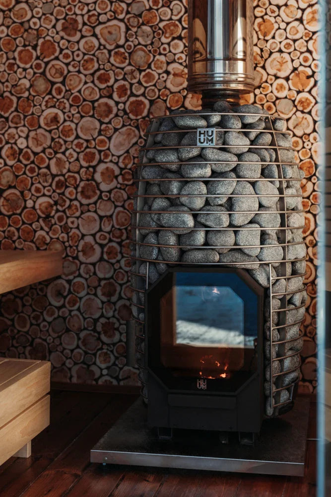 A HUUM Hive Flow LS Wood Burning Sauna Stove by HUUM, surrounded by sauna stones, stands on a wooden floor beside a log-patterned wall. A portion of a wooden bench is seen to the left.