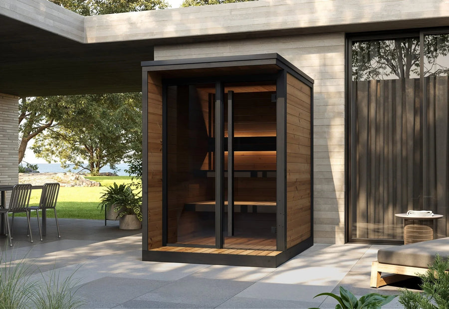 The SaunaLife Model GL4 4 Person Garden Luxury Series Outdoor Sauna Kit, with sleek glass doors, sits on a stylish patio outside a modern home. Potted plants, outdoor furniture, trees, and open space create an inviting atmosphere.