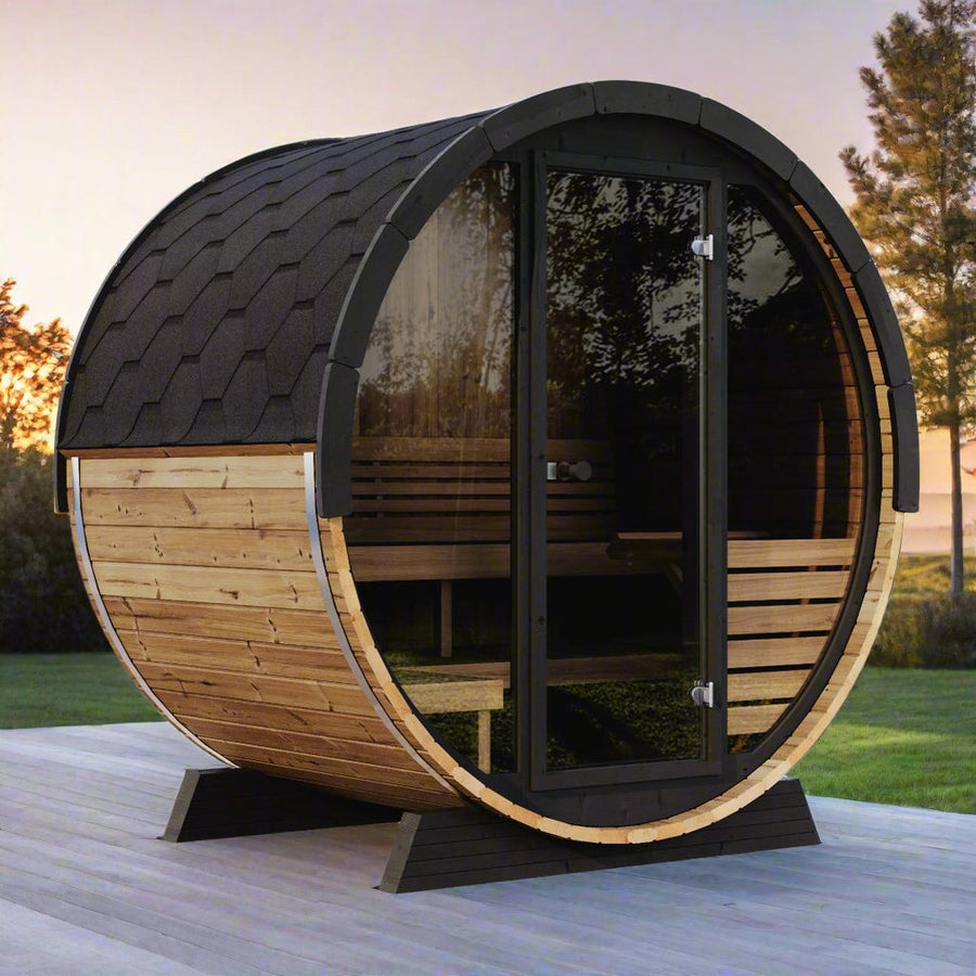 A SaunaLife Model EE6G 4 Person Barrel Sauna with a shingled roof and glass door by SaunaLife rests on a wooden deck outdoors at sunset, surrounded by trees and grass.