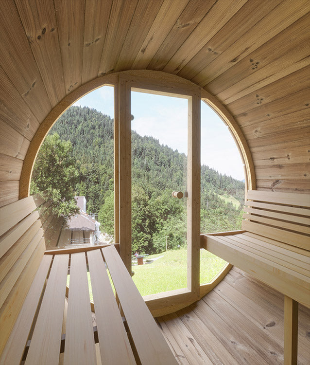 The SaunaLife 4-Person E7 ERGO Series Barrel Sauna by SaunaLife features side benches and a large circular window for enjoying views of green trees, grassy hills, and distant mountains in ultimate barrel sauna comfort.