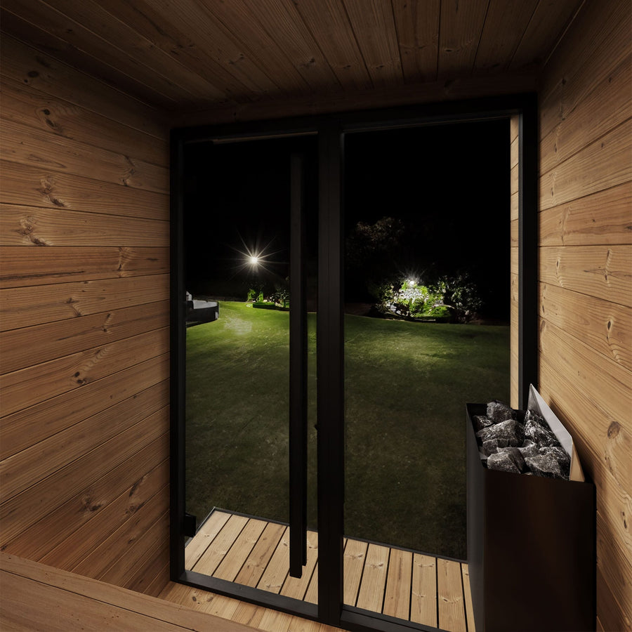Enjoy a nighttime view from the SaunaLife Model GL4 4 Person Garden Luxury Series Outdoor Sauna Kit, featuring a glass door to a softly lit backyard and a stone sauna heater—ideal for authentic Scandinavian design sauna experiences.