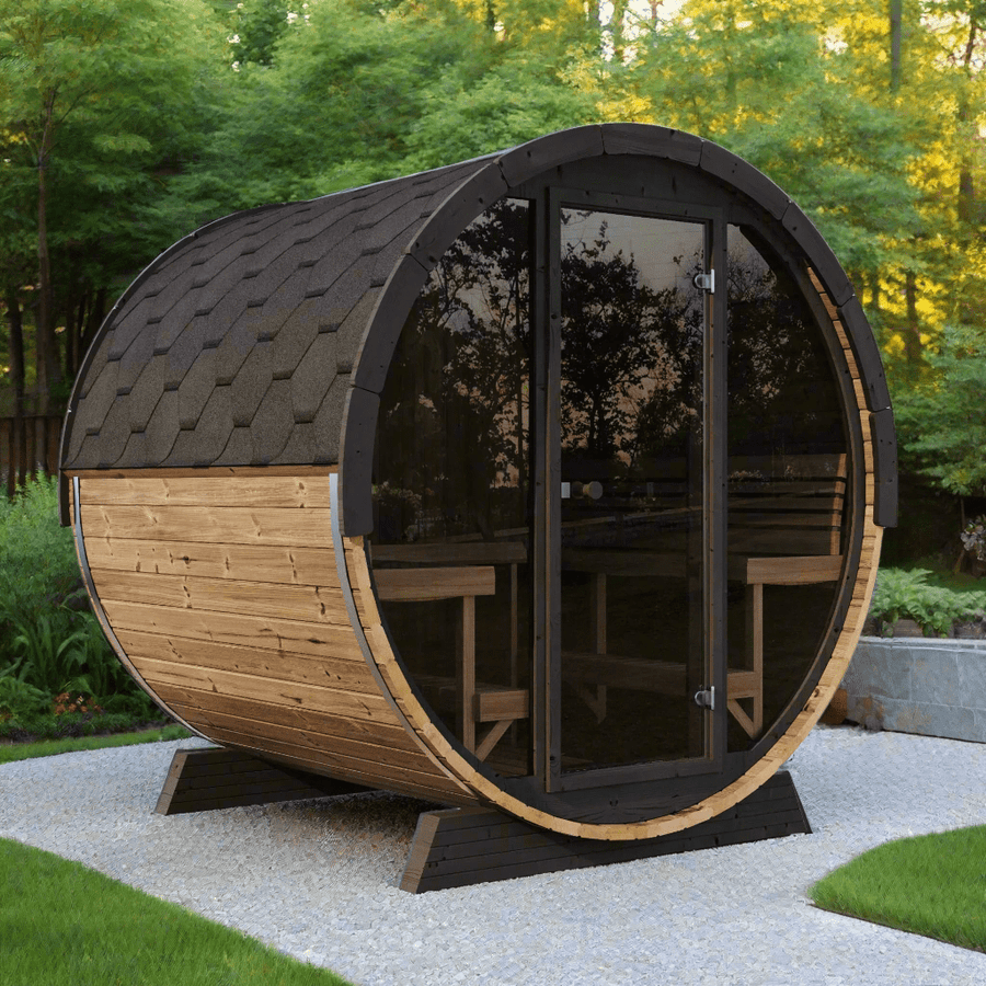 The SaunaLife Model EE8G 6 Person Barrel Sauna by SaunaLife, featuring a shingled roof, glass door, and interior benches, sits on a gravel patio amid green trees and lush backyard foliage.