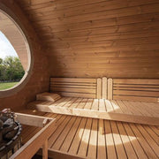 The SaunaLife Model G11 Outdoor 8 Person Sauna features a sloped ceiling, circular window, long bench with pillow, and a sauna heater with stones, all illuminated by sunlight highlighting the warm wood tones.