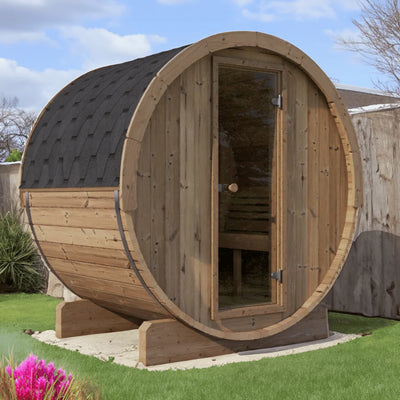 The SaunaLife Model E7W 4 Person Sauna Barrel-Window by SaunaLife features a shingled roof and glass door, sitting on a concrete pad in a grassy, fenced backyard—a stylish example of ergonomic outdoor sauna design.