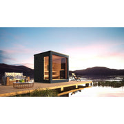 The SaunaLife G6 5-Person Outdoor Cabin Sauna by SaunaLife rests on a wooden deck beside a serene lake at sunset, with cozy seating and a lounge chair nearby, set against breathtaking mountain views.