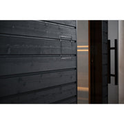 A close-up of the SaunaLife G6 5-Person Outdoor Cabin Sauna shows its modern black wooden wall with horizontal panels and a sleek metal handle by a glass door with a large black handle, softly lit from inside this stylish outdoor sauna by SaunaLife.
