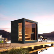 The SaunaLife G6 5-Person Outdoor Cabin Sauna features a modern black frame and large glass windows, set on a wooden deck over tranquil water at sunset, with mountains in the background, lounge chair, and table completing the serene scene.