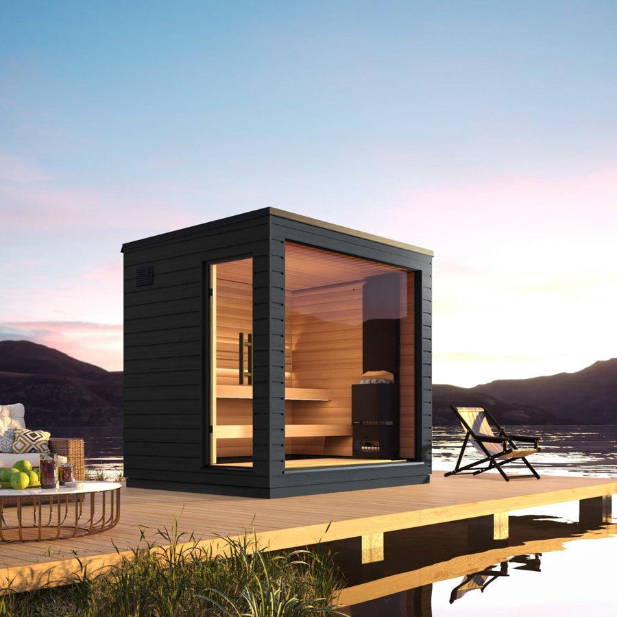 The SaunaLife G6 5-Person Outdoor Cabin Sauna features a modern black frame and large glass windows, set on a wooden deck over tranquil water at sunset, with mountains in the background, lounge chair, and table completing the serene scene.
