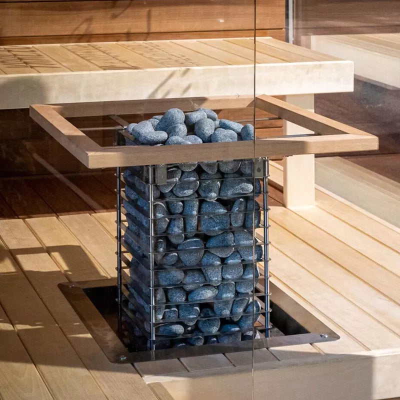 A modern sauna features a central HUUM CLIFF Series 11kW Sauna Heater Package by HUUM, filled with dark stones and surrounded by wooden benches and glass walls for powerful, efficient heating and a relaxing experience.