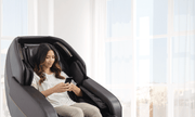 A woman relaxes in a Daiwa Hubble Massage Chair by Daiwa, enjoying an airbag massage while using her smartphone as sunlight streams through large windows, filling the room with natural light.
