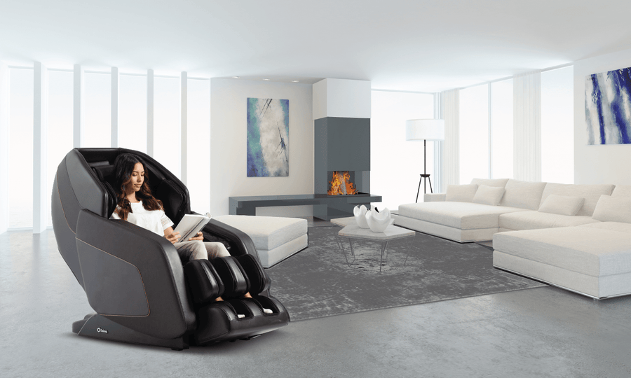 A woman unwinds in the Daiwa Hubble Massage Chair by Daiwa, enjoying a soothing airbag massage as she reads in a modern living room with white furniture, a gray rug, abstract art, and a glowing fireplace.