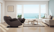 In a bright, minimalist living room with tall ocean-view windows, a person enjoys the heated compression leg massage of the Daiwa Legacy 4 Massage Chair by Daiwa, surrounded by a beige sofa, coffee table, and floor lamp.