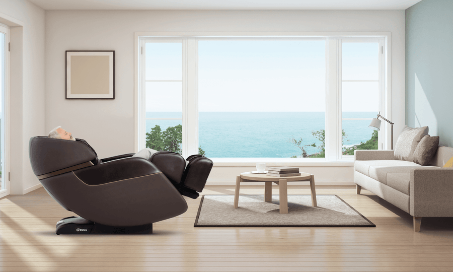 In a bright, minimalist living room with tall ocean-view windows, a person enjoys the heated compression leg massage of the Daiwa Legacy 4 Massage Chair by Daiwa, surrounded by a beige sofa, coffee table, and floor lamp.