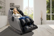 A woman relaxes in a Kyota Kansha M878 Massage Chair by Kyota, surrounded by a bright minimalist room with large windows, a vase of tall branches, and beautiful mountain views outside.