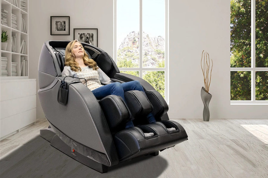 A woman relaxes in a Kyota Kansha M878 Massage Chair by Kyota, surrounded by a bright minimalist room with large windows, a vase of tall branches, and beautiful mountain views outside.