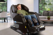 A man relaxes with his eyes closed in a Kyota Yosei M868 4D Massage Chair featuring lumbar heat, set in a modern living room with glass walls and greenery outside.