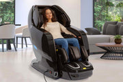 A woman relaxes in the Kyota Yosei M868 4D Massage Chair by Kyota, eyes closed and enjoying lumbar heat. The large black chair is set in a modern bright living room with a sofa, table, and plants in the background.