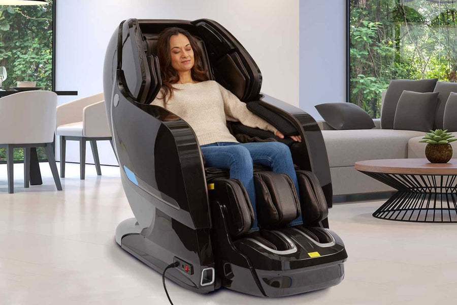 A woman relaxes in a Kyota Yosei M868 4D Massage Chair by Kyota, eyes closed and smiling as she enjoys a soothing foot massage, surrounded by light furniture and greenery seen through the windows.
