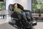 A man relaxes in the Kyota Yosei M868 4D Massage Chair by Kyota, featuring lumbar heat, in a modern living room with large windows that showcase lush greenery outside.