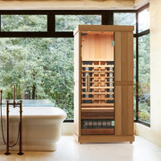 A modern bathroom with large windows features a standalone bathtub next to the Finnmark FD-1 Full-Spectrum Infrared Sauna by Finnmark, a stylish 1-person cabin with glass doors; lush green trees are visible outside.
