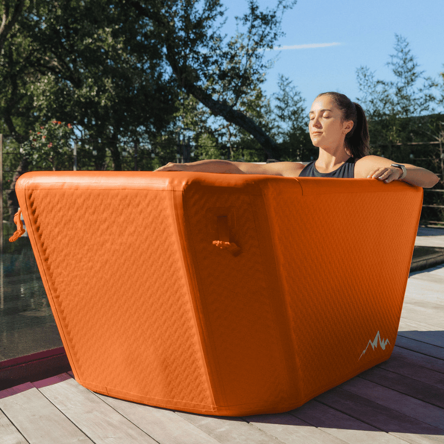 A woman relaxes with her eyes closed in The Big Tex Endurance Plunge Tub by Icebound Essentials, enjoying full-body immersion in the bright orange inflatable tub on a wooden deck surrounded by trees and sunlight.