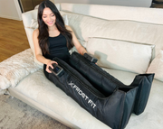 A woman smiles on a couch using Icebound Essentials Recovery Wraps™ Wireless Compression Boots, holding the controllers, with a plush blanket beside her for a cozy compression massage.