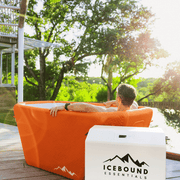 A person relaxes in The Big Tex Endurance Plunge + Chiller by Icebound Essentials on a deck by the lake, surrounded by trees. Nearby, a white Icebound Essentials cooler is ready for portable cold therapy.