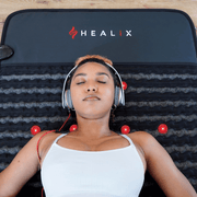 A woman in a white tank top lies relaxed on a HEALiX Infrared Revive PEMF Mat with red lights, eyes closed and wearing headphones. The mat, known for its red light therapy benefits, rests on a wooden floor.