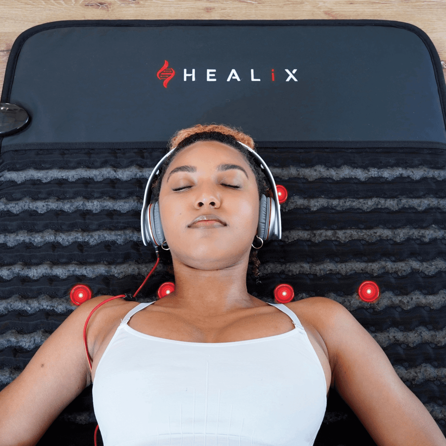 A woman in a white tank top lies relaxed on a HEALiX Infrared Revive PEMF Mat with red lights, eyes closed and wearing headphones. The mat, known for its red light therapy benefits, rests on a wooden floor.