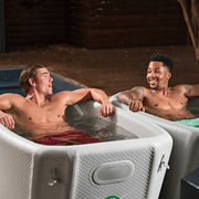 Two men relax in separate Icebound Essentials Endurance Plunge Tubs, partially submerged and smiling as they chat together indoors or in a covered outdoor space.