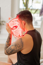 A person with a tattooed arm and short hair holds the Mito Red Light MitoGLOW™ LED Red Light Therapy Mask—with blue, yellow, red, and near infrared LEDs—up to their face indoors while wearing a sleeveless black top.