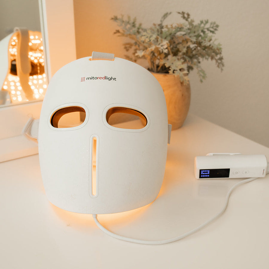 The MitoGLOW™ LED Red Light Therapy Mask by Mito Red Light, featuring blue, yellow, red, and near infrared LEDs for acne treatment, rests on a white table connected to its control device near a mirror and potted plant.