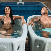 A woman in a black bikini and a man in light swim shorts relax separately in two Icebound Essentials Endurance Plunge + Chiller ice baths, filled with water and set side by side next to an outdoor pool.