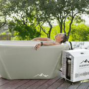 A person relaxes in The Endurance Plunge + Chiller by Icebound Essentials, surrounded by greenery and mountains, enjoying the benefits of cold therapy next to the white chiller unit.