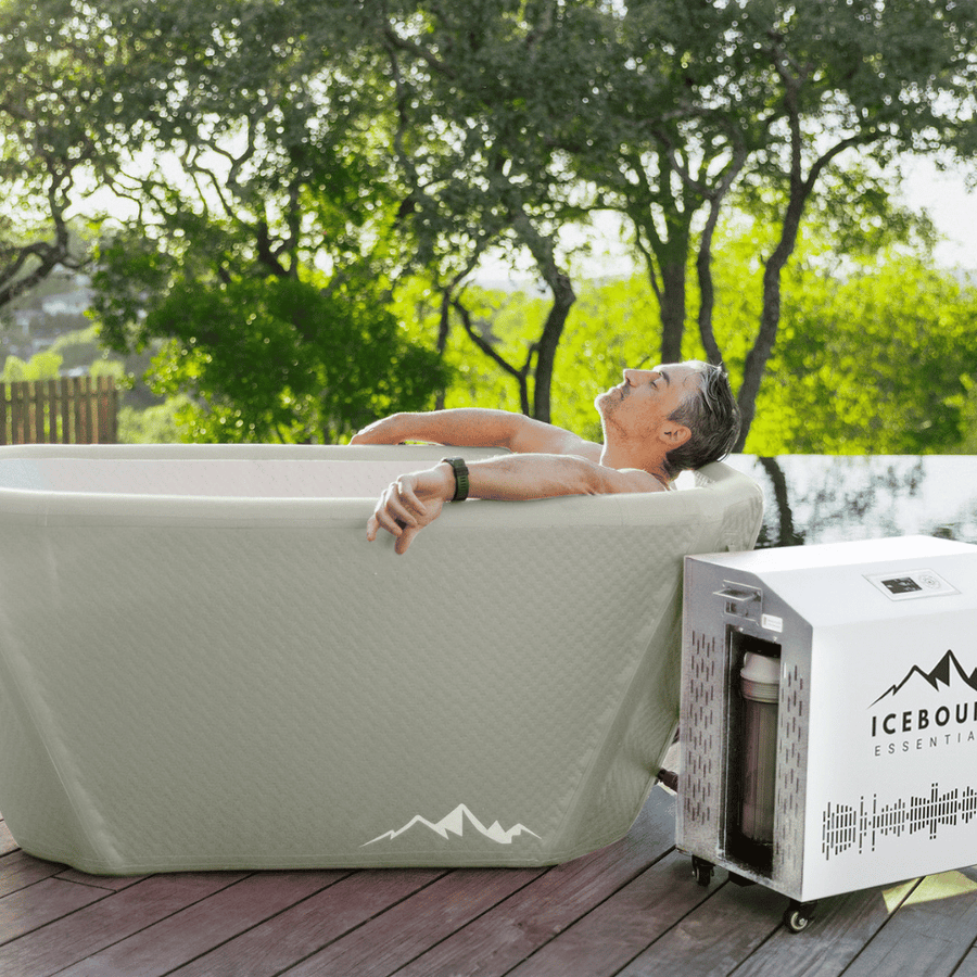 A person relaxes in The Endurance Plunge + Chiller by Icebound Essentials, surrounded by greenery and mountains, enjoying the benefits of cold therapy next to the white chiller unit.