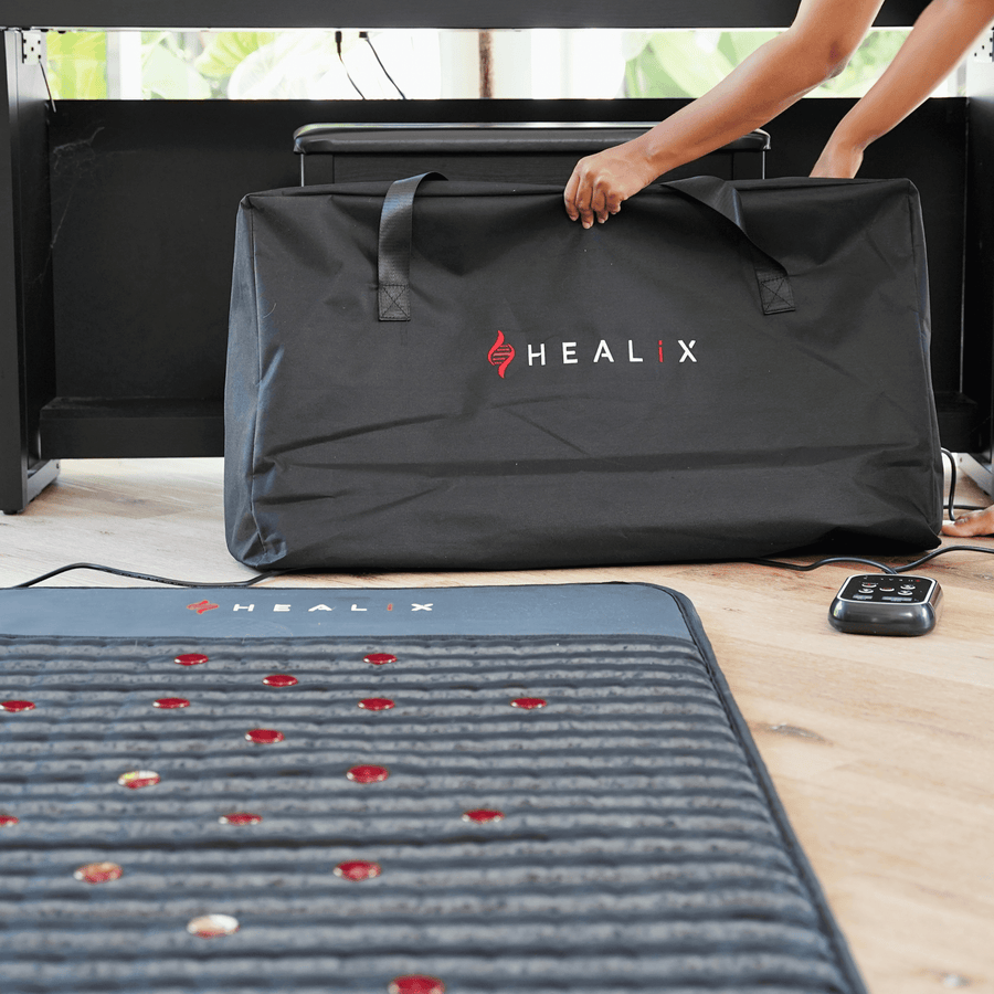 A person sets up a HEALiX Revive mat from HEALiX Infrared on a wooden floor beside its black carrying case and remote control. The mat features embedded gemstones and red lights for red light and soothing infrared therapy.