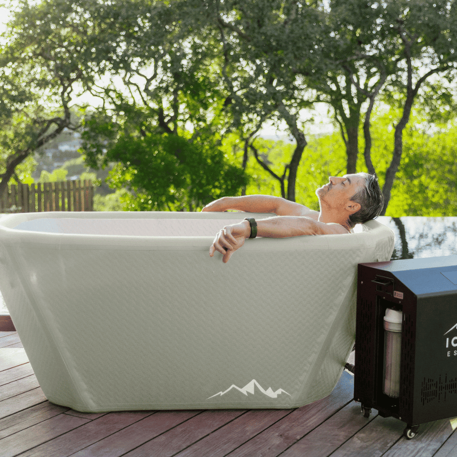 A man relaxes in The Endurance Plunge + Chiller by Icebound Essentials on a wooden deck surrounded by trees, with the black chiller unit nearby and sunlight filtering through the leaves.
