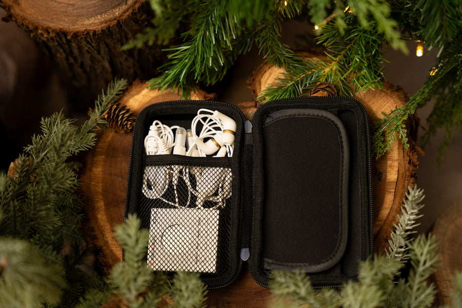 The MitoBOOST™ Intranasal Device System by Mito Red Light, for sinus, immune, and brain health, comes in a black zippered carrying case with white earbuds and a charging cable, displayed on wooden logs among green pine branches.