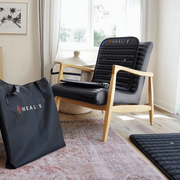 A cozy room with a wooden armchair featuring a HEALiX Infrared-branded black cushion and backrest. A HEALiX Revive Mini PEMF Mat lies on the floor, promoting relaxation, while a large HEALiX carrying bag rests nearby in streamed sunlight.