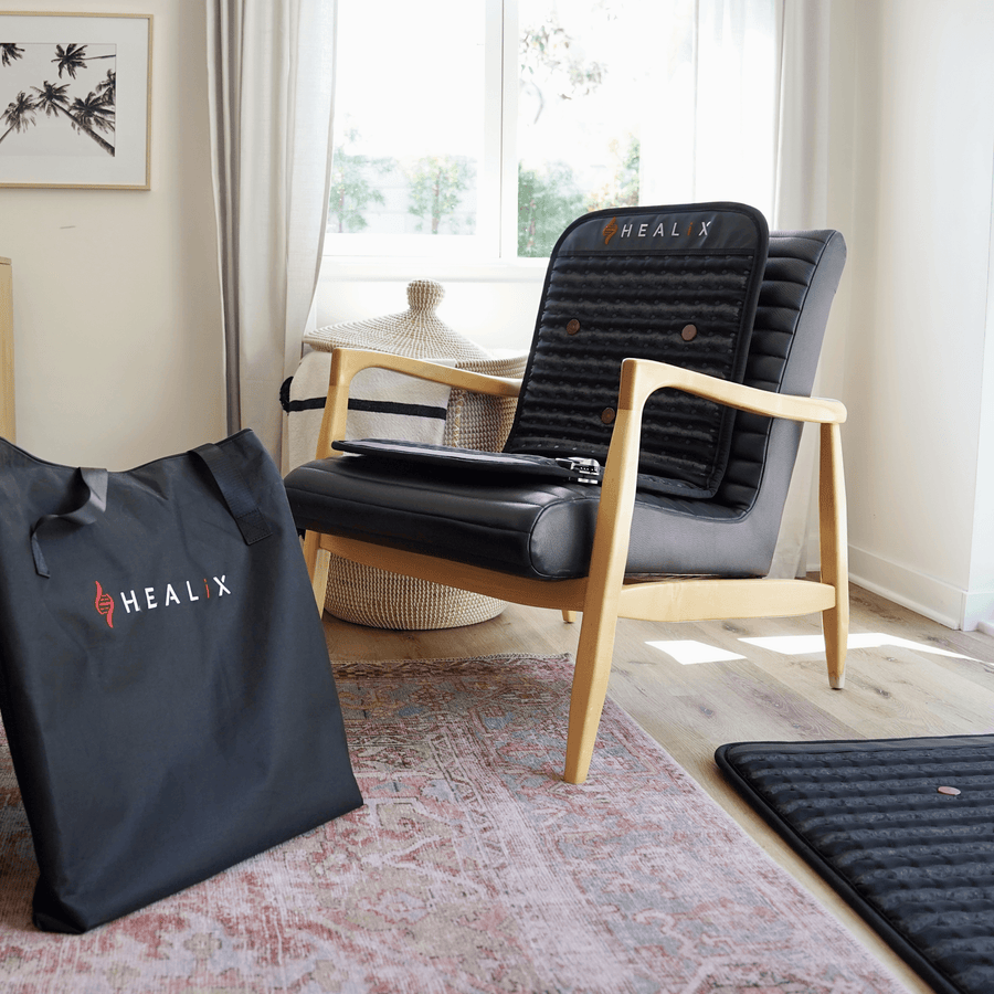 A cozy room with a wooden armchair featuring a HEALiX Infrared-branded black cushion and backrest. A HEALiX Revive Mini PEMF Mat lies on the floor, promoting relaxation, while a large HEALiX carrying bag rests nearby in streamed sunlight.