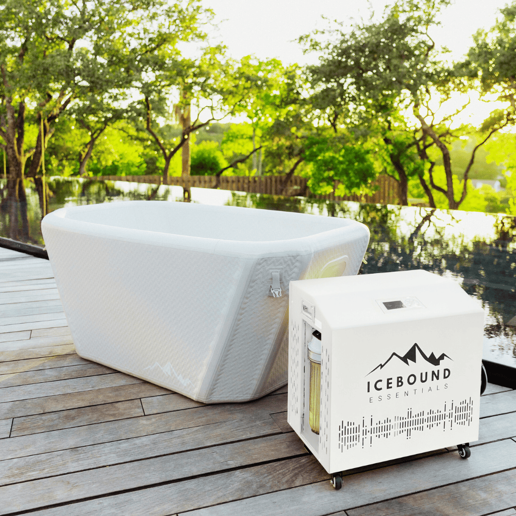 An Icebound Essentials Endurance Plunge + Chiller is set up on a wooden deck beside a reflective pool, providing portable cold therapy amid green trees and bright sunlight.