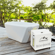 The Endurance Plunge + Chiller by Icebound Essentials, a white rectangular inflatable ice bath with matching cooling unit, sits on a wooden deck by a sleek pool—lush green trees and bright sunlight create the perfect cold therapy setting.