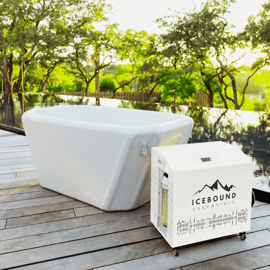 The Endurance Plunge + Chiller by Icebound Essentials, a white rectangular inflatable ice bath with matching cooling unit, sits on a wooden deck by a sleek pool—lush green trees and bright sunlight create the perfect cold therapy setting.