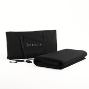A black HEALiX Infrared carrying case stands upright beside a rolled-up black mat—the HEALiX Glow Pod—with its control remote and power cord displayed in front, all set against a white background.