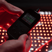 A person holds a handheld Elevate Light Pod by HEALiX Infrared, showing “1658” and red control buttons, above a surface of glowing red LED lights for pain relief and red light therapy.