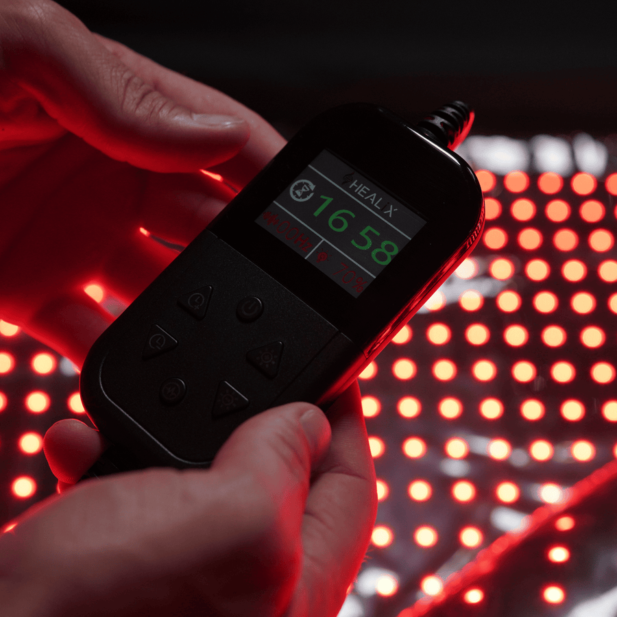 A person holds a handheld Elevate Light Pod by HEALiX Infrared, showing “1658” and red control buttons, above a surface of glowing red LED lights for pain relief and red light therapy.