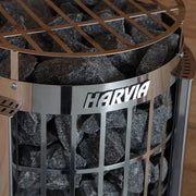 Close-up of the Harvia Cilindro PC90E 240V sauna heater with external controls, featuring a grid-like exterior filled with dark stones. The HARVIA brand name stands out against light wooden paneling in the background.