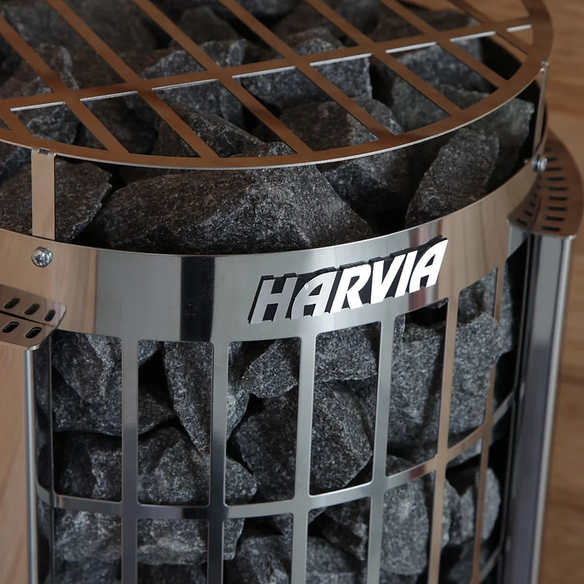 Close-up of the Harvia Cilindro PC90E 240V sauna heater with external controls, featuring a grid-like exterior filled with dark stones. The HARVIA brand name stands out against light wooden paneling in the background.