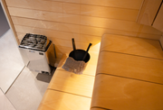 A cozy Finnish sauna room features light wood benches, a black water ladle and bowl, a gray towel, and a Harvia KIP45B Stainless Steel 4.5KW Sauna Heater with Built-in Control by Harvia in the corner for a warm, inviting atmosphere.
